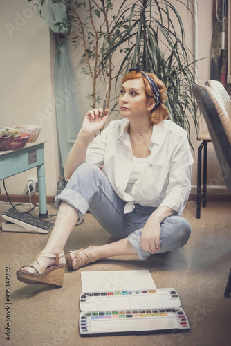 A young active female artist, with red hair, in a white shirt and jeans with a bandage on her head, paints standing at the table photo