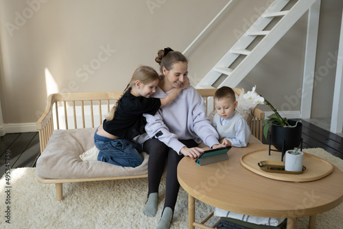 Mom works. A happy daughter and son hug their mom on the couch in the room and watch their mother working on a tablet. Internet and online technologies. Remote work on maternity leave. © Lys Owl