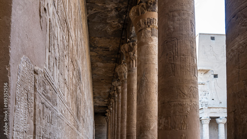 Colonnade in the Temple of Horus in Edfu. There is a narrow passage between a row of columns with capitals and the wall. Hieroglyphs and carvings are visible. Egypt.