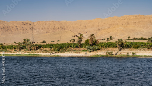 Palm trees and green vegetation grow on the banks of the Nile. Power towers are visible. A high sand dune against a clear sky. Ripples on the blue water. Egypt