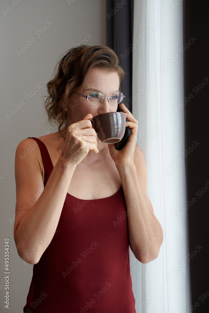 Woman in silk burgundy dress drinking cup of delicious coffee and looking through window and talking on phone Woman in silk burgundy dress drinking cup of delicious coffee and looking through window and talking on phone