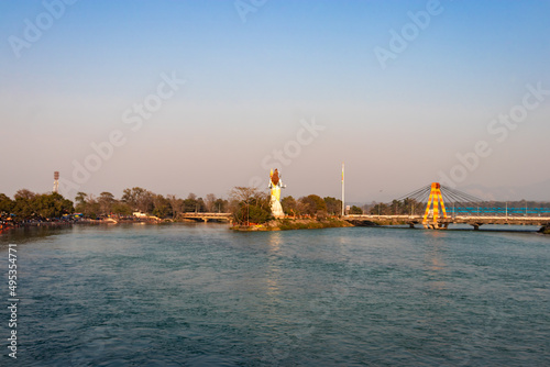 hindu god shiva statue and isolated cable bridge over ganges river from back with bright blue sky
