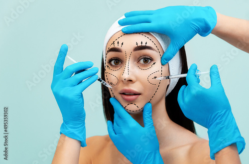 Portrait of a pretty young woman with surgical markings on her face. Hands of doctors in medical gloves lean syringes against a woman s face. Concept of cosmetic injections