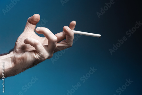 Fototapeta Naklejka Na Ścianę i Meble -  World No Tabacco day. A pale, crooked male hand, close-up, holding a smoking cigarette. Dark blue background. Copy space. The concept of nicotine addiction