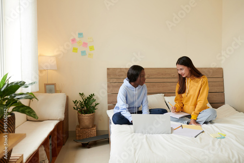 Smiling young woman explaining difficult topic to friend when they are sitting on bed in dormitory photo