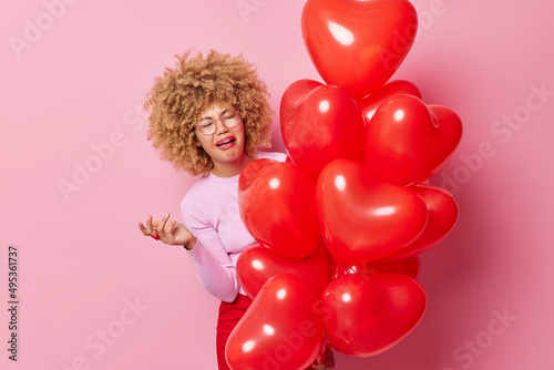 Fotótapéta Upset crying woman has spoiled makeup curly hair feels dejected holds bunch of heart balloons keeps hand raised expresses negative emotions isolated over pink background