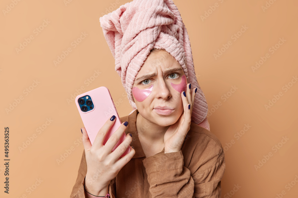 Photo of displeased young upset woman has healthy skin applies beauty hydrogel patches dressed in nightwear wears towel on head after taking bath uses cellphone poses against brown background Photo of displeased young upset woman has healthy skin applies beauty hydrogel patches dressed in nightwear wears towel on head after taking bath uses cellphone poses against brown background
