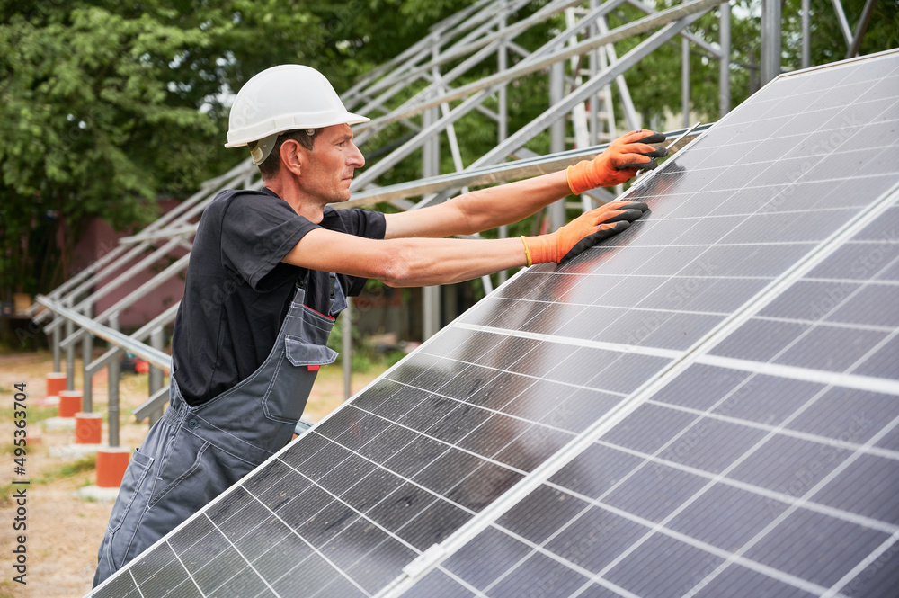 Man in safety construction helmet mounting photovoltaic solar panel. Male technician in work gloves assembling solar modules, tightening with hex key. Concept of alternative energy sources.