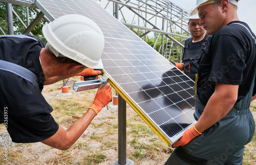 Male workers mounting photovoltaic solar panel system outdoors. Three men wearing safety helmets and work overalls while placing solar module on metal supporting structure. © anatoliy_gleb