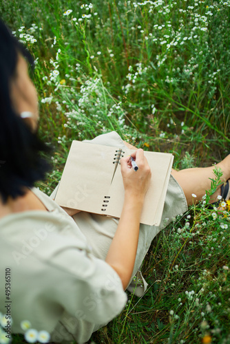 Female woman with pen writing or painting, handwriting on notebook on flower blooming meadow
