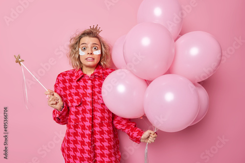 Scared terrified woman holds magic wand and bunch of inflated balloons cannot believe her eyes feels fearful applies beauty patches under eyes stands against pink background. Birthday party. © wayhome.studio 