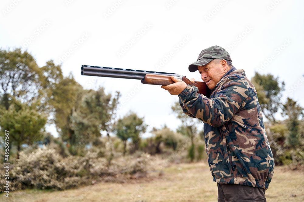 Fototapeta premium Senior male hunter in camouflage clothing in the field aiming hunting shotgun during a hunt. High quality photo