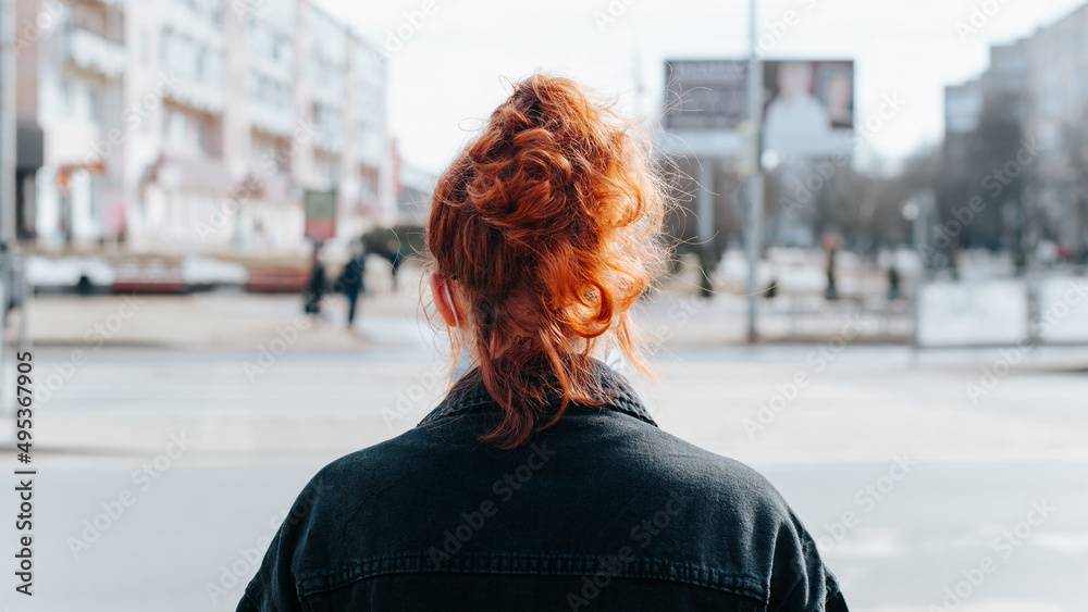 Back view redhead curly young woman standing street bokeh, unrecognizable girl pedestrian outdoors during daytime, closeup