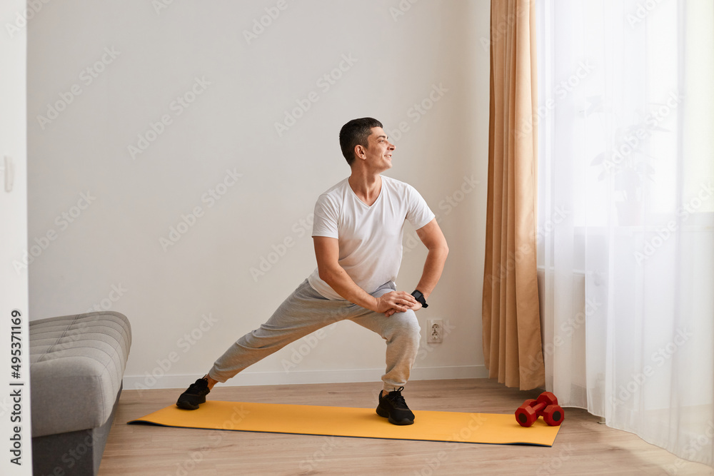 Indoor shot of man wearing sportswear doing sport exercises at home, doing leg stretching exercises on mat before workout, healthy lifestyle, practicing yoga, looking away. Indoor shot of man wearing sportswear doing sport exercises at home, doing leg stretching exercises on mat before workout, healthy lifestyle, practicing yoga, looking away.