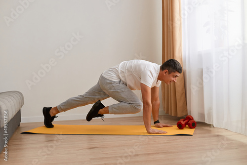 Side view full length portrait of man wearing sportswear doing sport exercises at home, practicing yoga, doing plank bent leg, training strength and muscle flexibility.