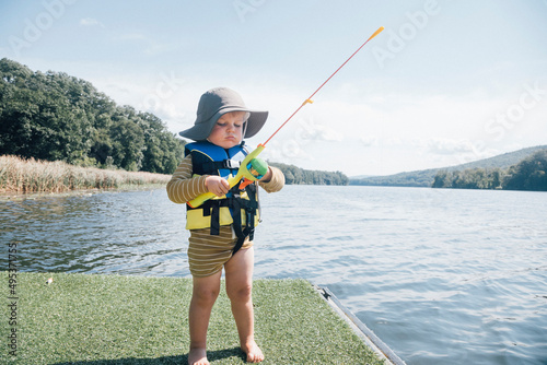 Cute baby boy with fishing rod standing by river on sunny day