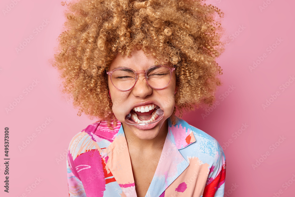 Headshot of curly haired woman with expander in mouth shows white teeth keeps eyes closed wears spectacles and colorful shirt poses against pink background. Emotional female uses dental retractor Headshot of curly haired woman with expander in mouth shows white teeth keeps eyes closed wears spectacles and colorful shirt poses against pink background. Emotional female uses dental retractor