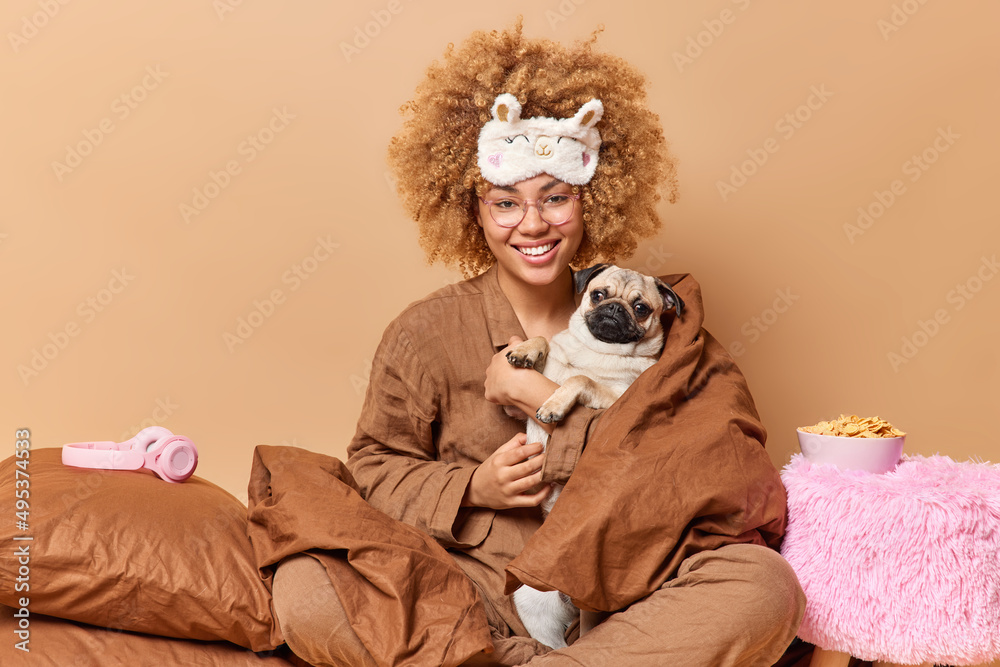 Obraz premium Happy female pet owner with cute pug dog wrapped in blanket wears pajama and sleepmask plays with favorite pet pose in bedroom against brown background. Domestic atmosphere animals friendship