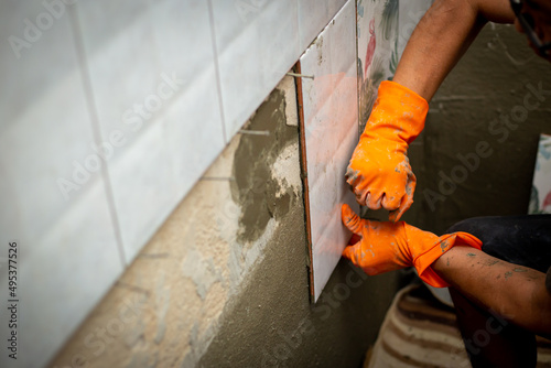 Builder tiling a concrete wall with decorative ornamental tiles lining up a tile with his gloved hands to seat into the tiling cement on the wall. © WITTAYA