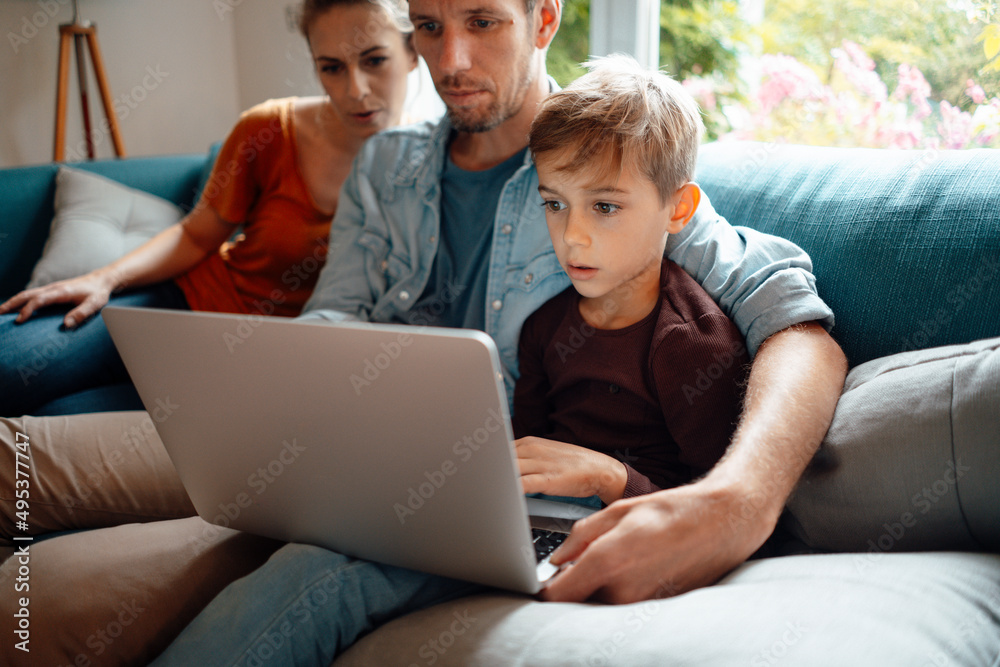 Father and son sharing laptop sitting by woman on sofa at home Father and son sharing laptop sitting by woman on sofa at home
