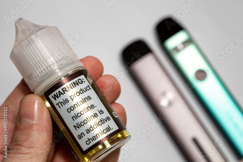 Electronic cigarette liquid bottle selective focus. On the plastic bottle it says "Warning: This product contains nicotine. Nicotine is an addictive chemical." Two nicotine vapor sticks, out of focus.
