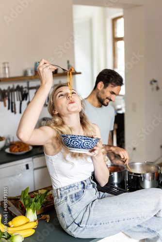 Woman slurping noodles with boyfriend preparing food in kitchen at home photo