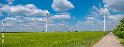 Panoramic view over beautiful farm landscape with green wheat field, lonely road and wind turbines to produce green energy in Germany, Spring, at blue sky and sunny day.