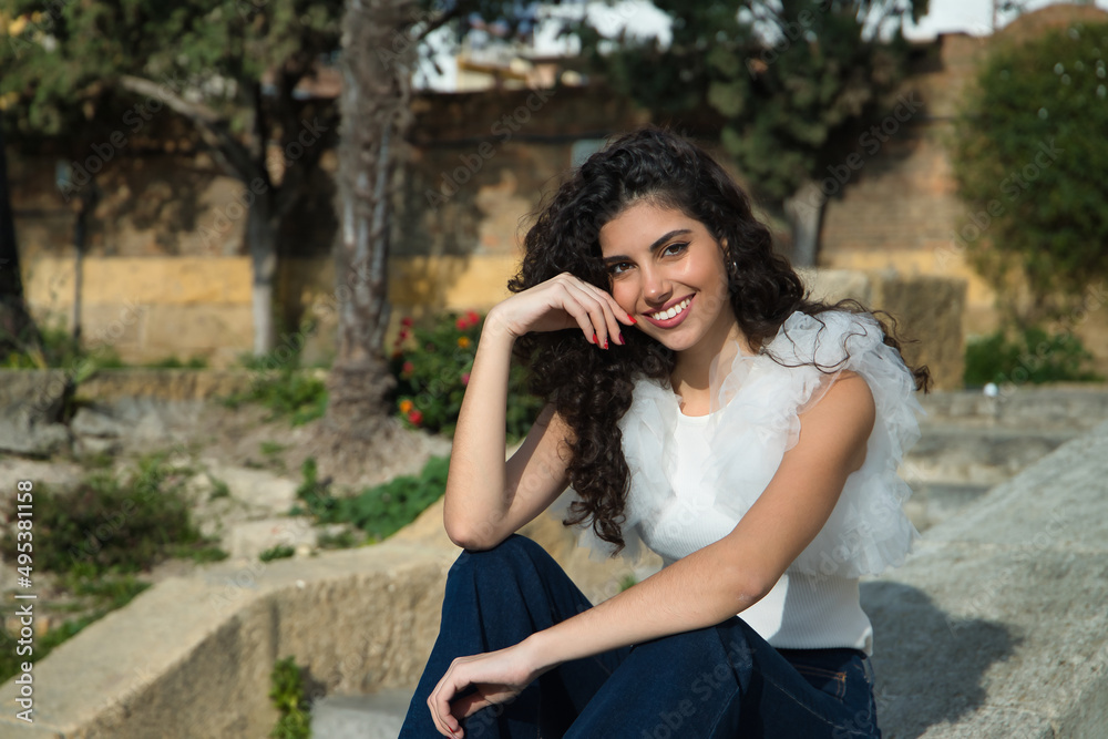 Young and beautiful woman, brunette, with curly hair and a nice smile, wearing a white shirt and jeans, sitting on a concrete wall. Concept beauty, fashion, model, happiness, trend. Young and beautiful woman, brunette, with curly hair and a nice smile, wearing a white shirt and jeans, sitting on a concrete wall. Concept beauty, fashion, model, happiness, trend.