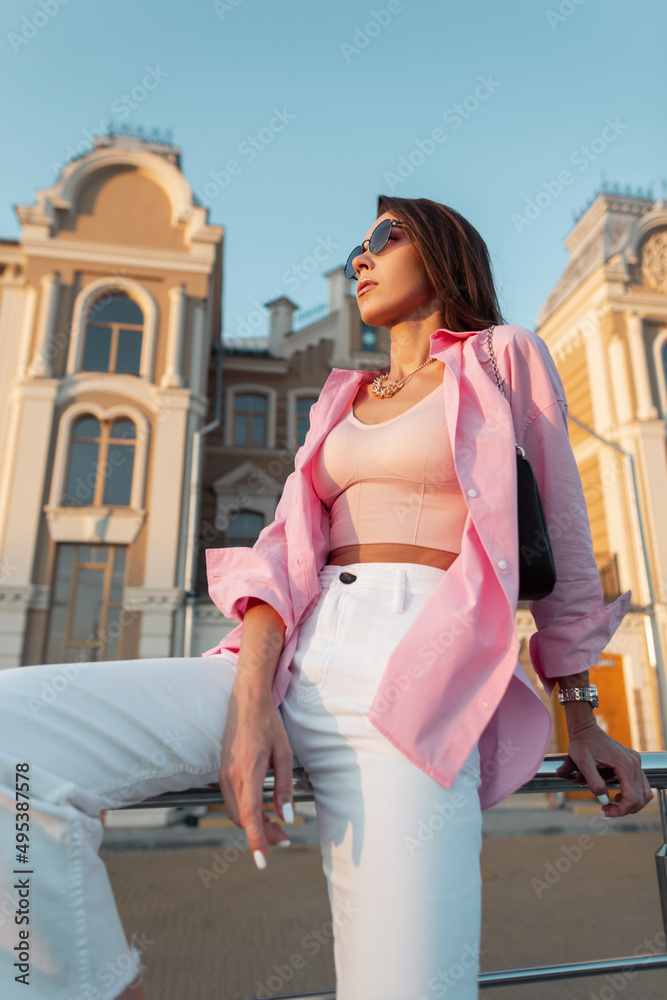 Trendy fashion young woman model with stylish glasses in trendy pink shirt, tank top, white jeans and handbag sits in the city. Summer urban female style look