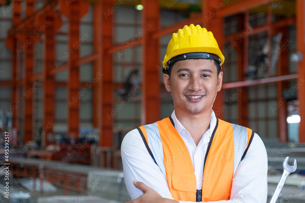 Selective focus at Asian men mechanical engineer, wearing safety equipment. While doing audit for quality and safety control inside of factory area. With blurred background of heavy machine.