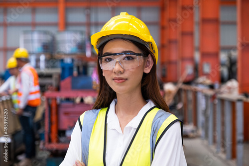 Selective focus at young women inspector wearing safety equipment. While doing audit for quality and safety control inside of factory area. With blurred background of heavy machine.