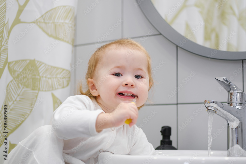 Close up of cute toddler sitting on the washbasin cabinet and brushing teeth, showing independence, learning new skills. Daily morning hygiene routine. Personal and teeth hygiene. Close up of cute toddler sitting on the washbasin cabinet and brushing teeth, showing independence, learning new skills. Daily morning hygiene routine. Personal and teeth hygiene.