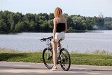 Slim redhead girl in shorts stopped with bicycle on river and forest background. Cycling and summer leisure