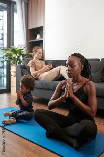 Young woman and her toddler son meditating sitting in lotus position on yoga mat at home