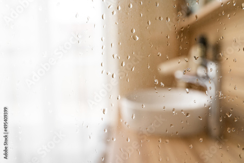 Water drops on wet glass shower door in hotel bathroom with blured bokeh window, sink, faucet and wooden furniture on sunny morning or day. Travel, holiday, vacation, interior design, body care