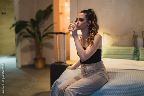 Chica delgada con pelo largo bebiendo agua en vaso de cristal al borde de una cama de hotel