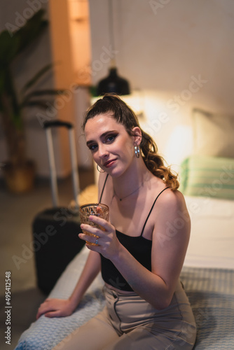 Chica delgada con pelo largo bebiendo agua en vaso de cristal al borde de una cama de hotel