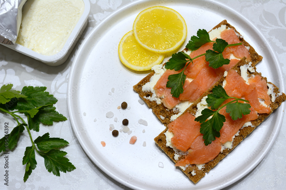 crispbread with soft cheese, salmon slices and parsley on white plate with lemon slices and allspice crispbread with soft cheese, salmon slices and parsley on white plate with lemon slices and allspice