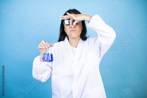 Young brunette woman wearing scientist uniform holding test tube over isolated blue background smelling something stinky and disgusting, intolerable smell, holding breath with fingers on nose