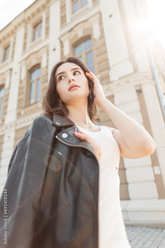 Caucasian beautiful model girl in fashionable summer clothes with black leather jacket and white dress walking in the city