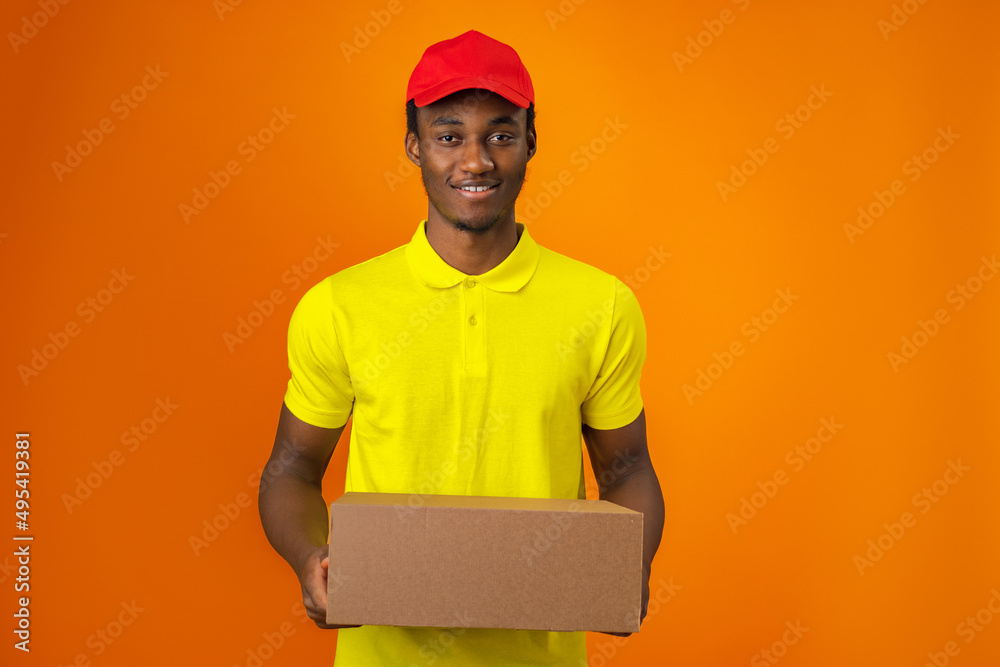 Smiling black male courier wearing uniform holding box in orange studio