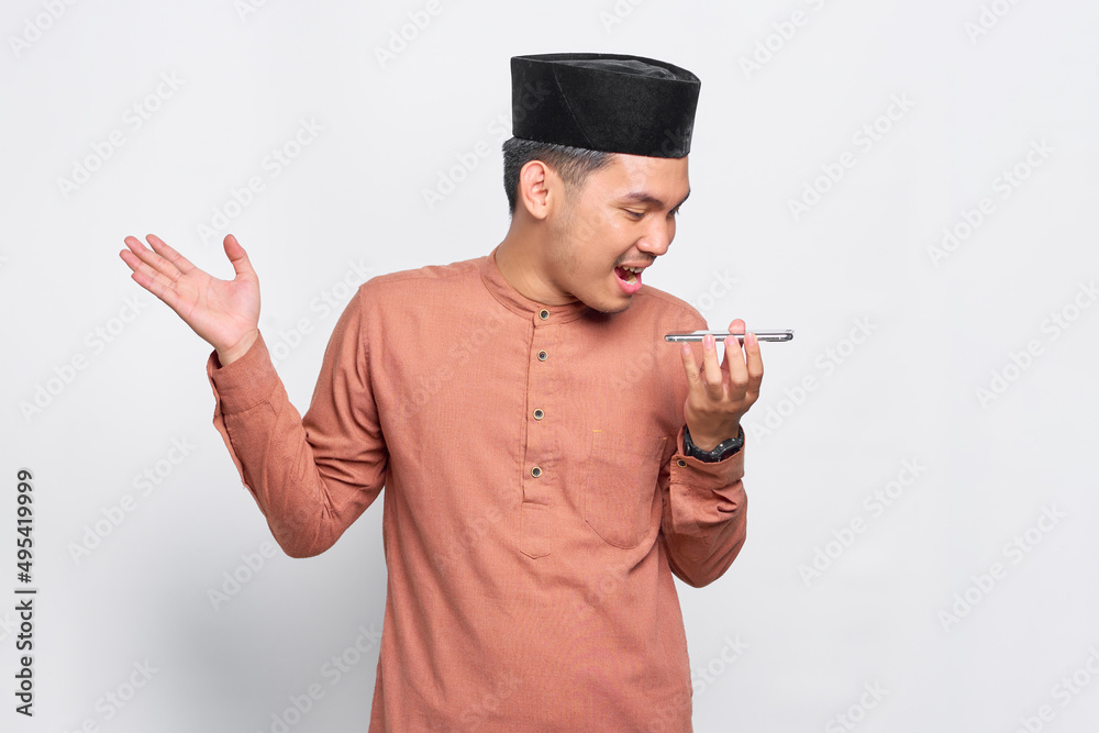 Cheerful young Asian Muslim man using the voice assistant on mobile phone isolated over white background