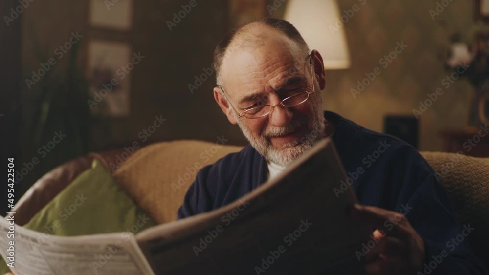 Cheerful senior man sitting on the couch in living room and laughing while watching latest news in the newspaper and reading jokes Cheerful senior man sitting on the couch in living room and laughing while watching latest news in the newspaper and reading jokes