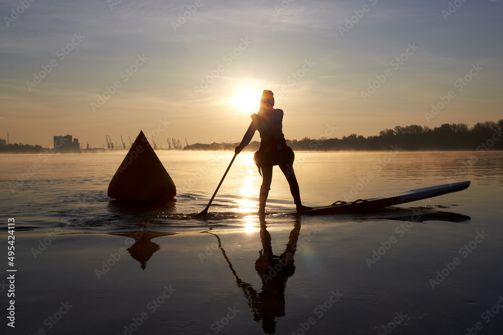 Silhouette of a young woman paddle on stand up paddle boarding (SUP) near inflatable buoy at sunrise on quiet autumn Danube river. Calm river surface at the morning. Morning training and meditation