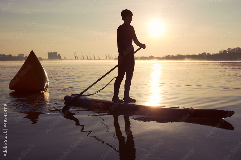 Silhouette of a young woman paddle on stand up paddle boarding (SUP) near buoy at sunrise on quiet autumn Danube river. Calm river surface at the morning. Morning training and meditation on the water