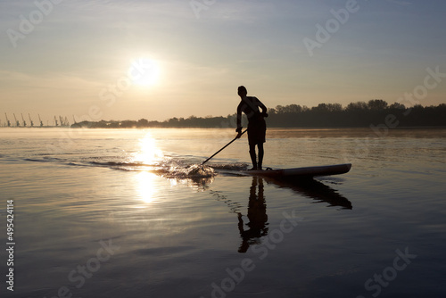 River landscape with silhouette of a man paddle on stand up paddle boarding (SUP) at sunrise on quiet surface of autumn Danube river. Morning training and meditation on the water