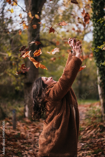 Medium shot of a brunette woman throwing dry leaves into the air in a forest.