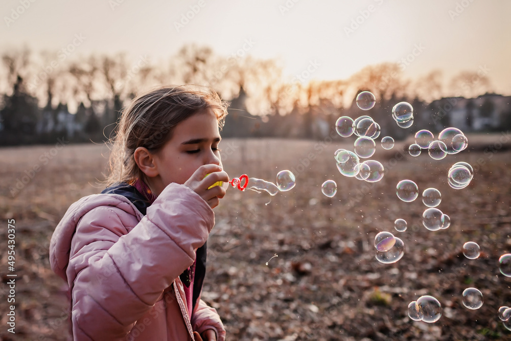 Ten years pretty girl in light pink coat blowing soap bubbles in forest at sunset, warm walking in early spring. Active leisure, healthy lifestyle. Happy peaceful childhood. Backlit evening sunshine
