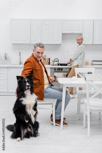 Mature man petting border collie near cup of coffee and blurred wife in kitchen.