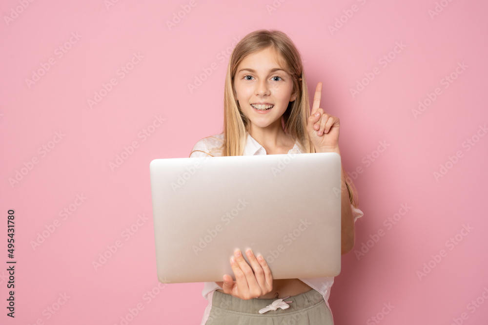 Portrait of little girl holding laptop computer while standing and looking at camera pointing finger up isolated over pink background Portrait of little girl holding laptop computer while standing and looking at camera pointing finger up isolated over pink background